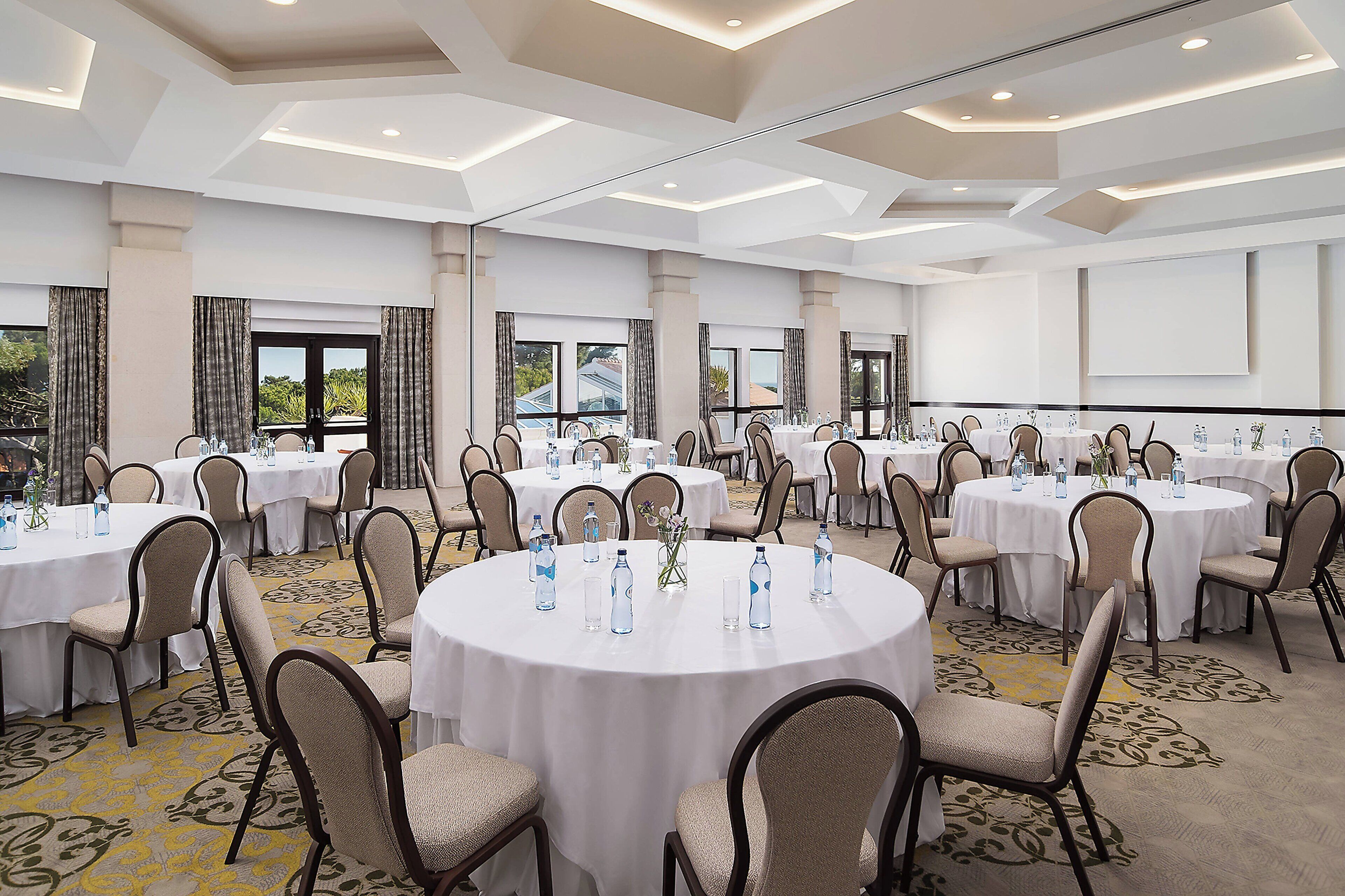 A conference room with multiple round tables, each surrounded by chairs, and large windows providing natural light.