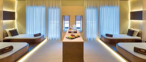 A hotel room with two beds, a central table with fruits, and a view through large windows.