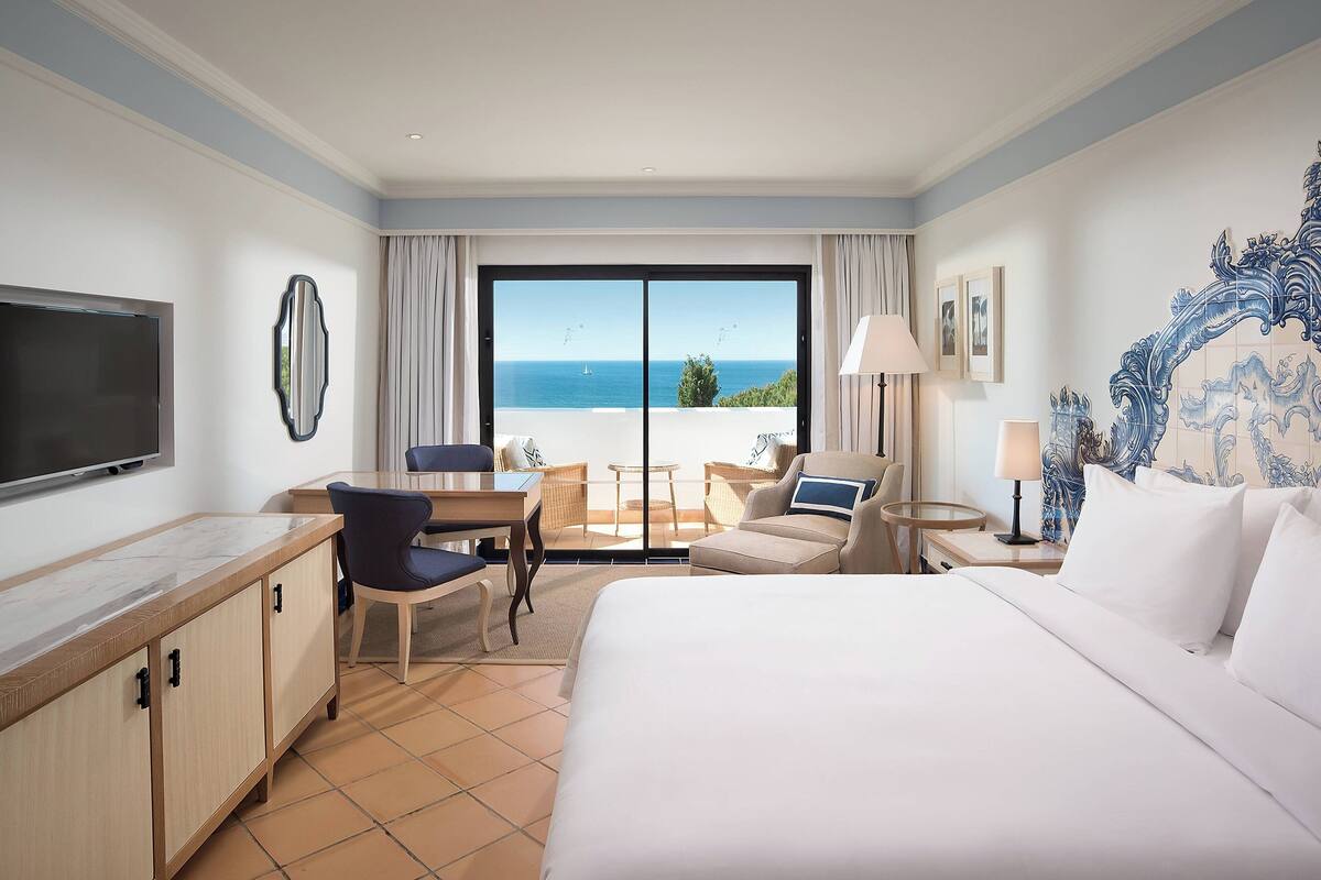 A hotel room with a bed, a desk, a chair, a TV, and a view of the sea.