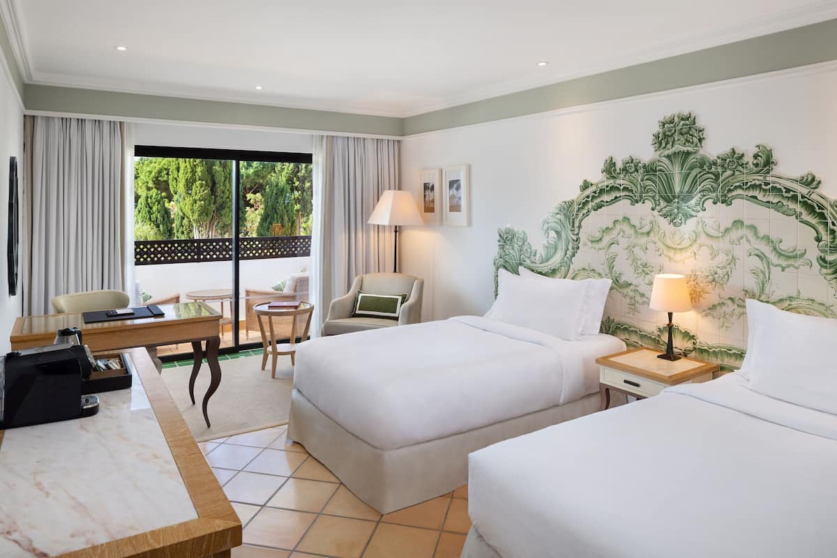 A hotel room with two beds, a desk, a chair, a lamp, and a view of greenery outside.