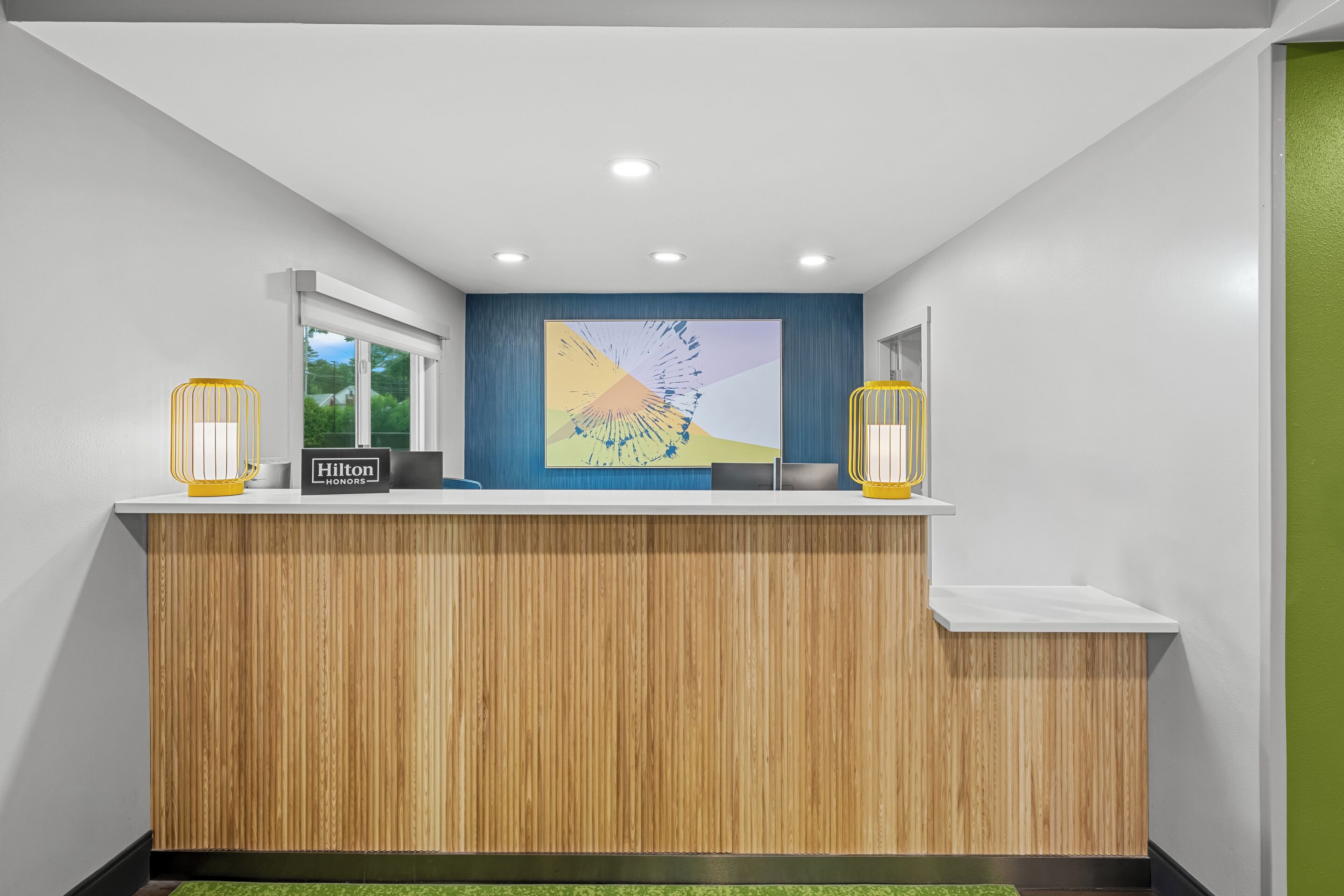 Un mostrador de recepción de hotel con encimera de madera, un cuadro abstracto grande y dos lámparas amarillas.