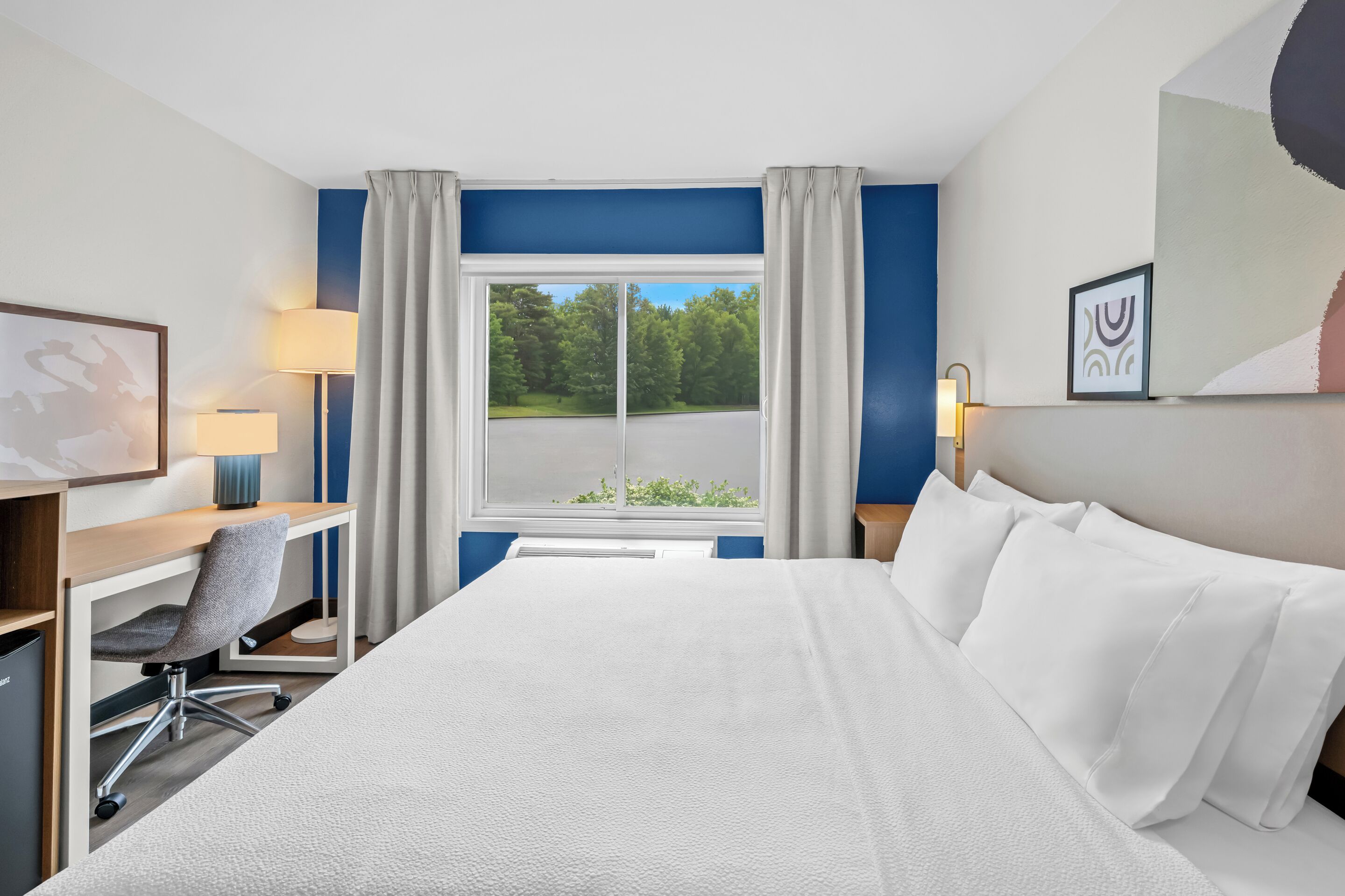 A hotel room with a bed, desk, chair, lamp, and a window with a view of trees.