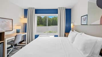 A hotel room with a bed, desk, chair, lamp, and a window with a view of trees.