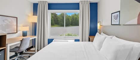 Une chambre d’hôtel équipée d’un lit, d’un bureau, d’une chaise, d’une lampe et d’une fenêtre donnant sur des arbres.