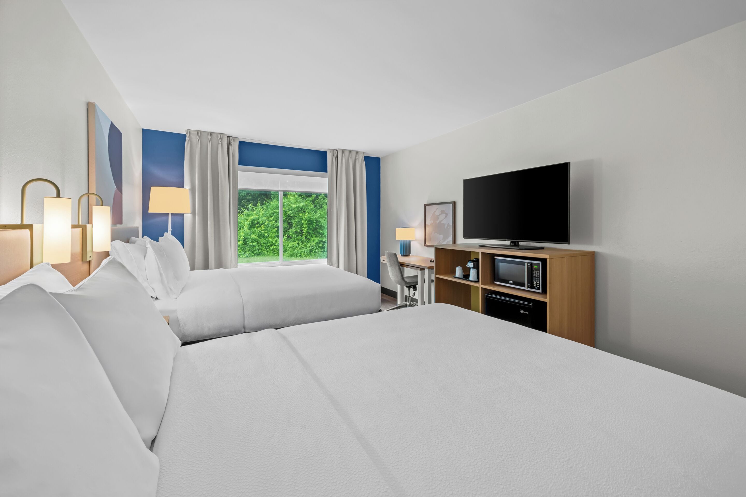 A hotel room with two beds, a television, a desk with a chair, a microwave, and a window with curtains.