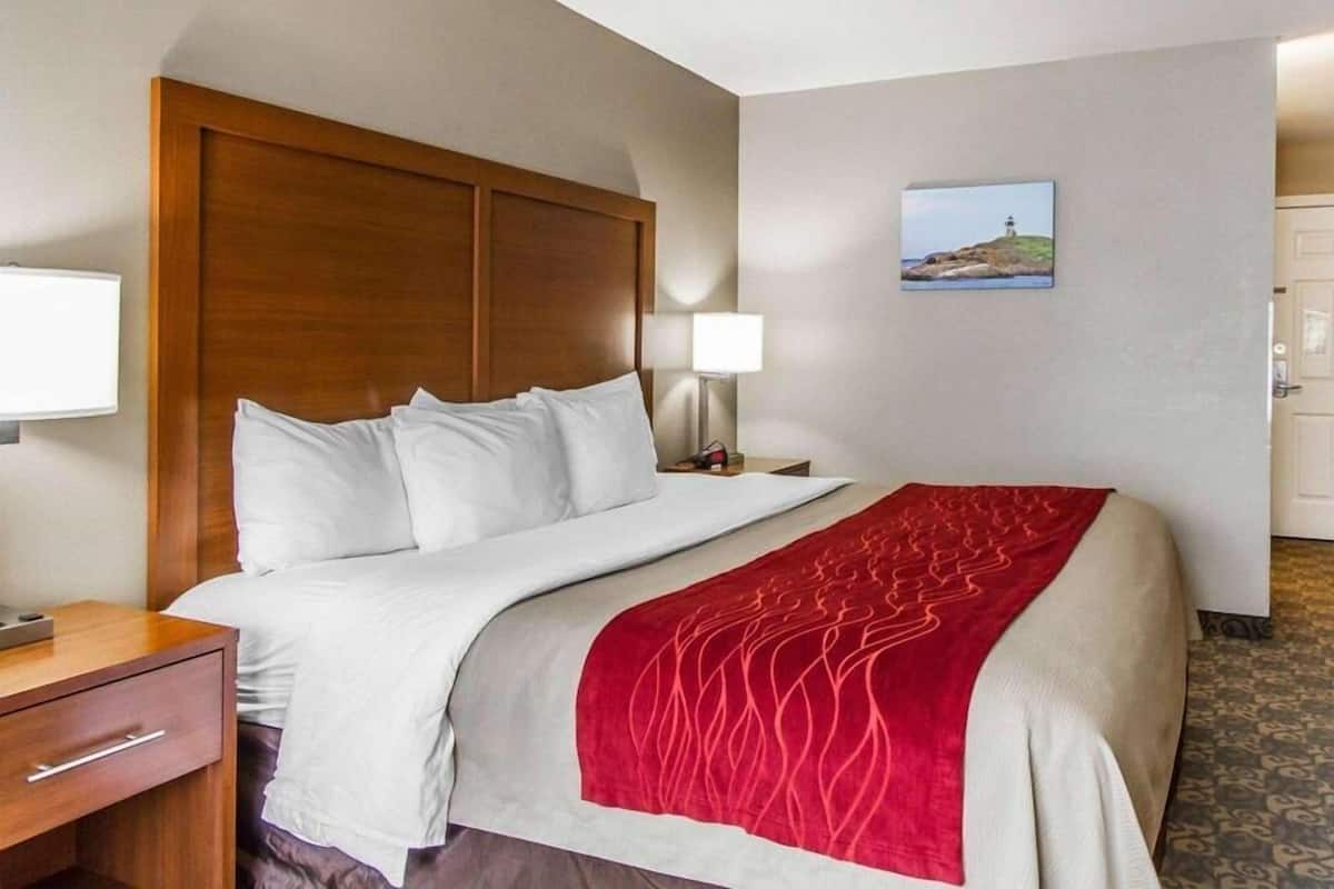 A hotel room with a wooden headboard, a bed with white bedding and a red accent throw, a nightstand with a lamp, and a painting of a lighthouse on the wall.
