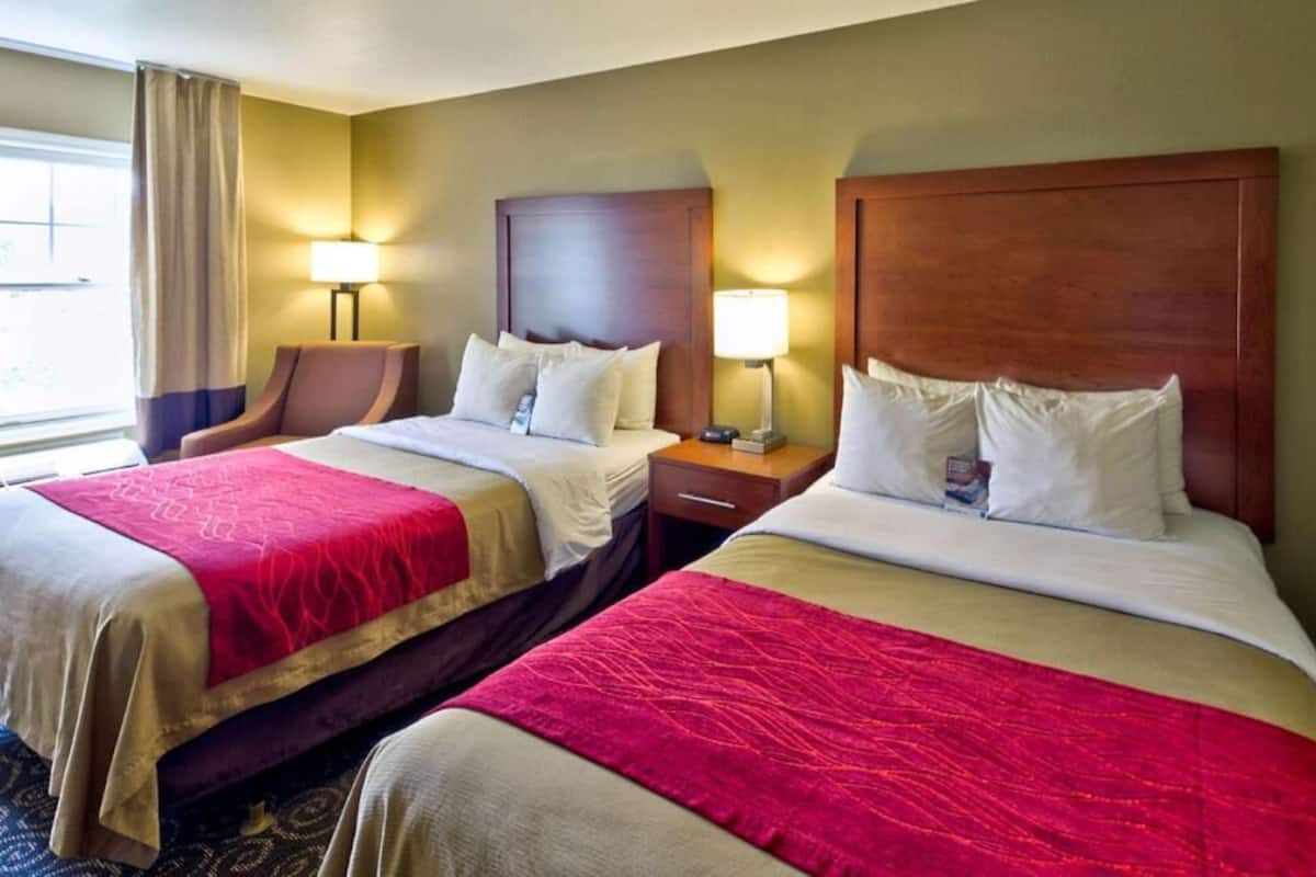 A hotel room with two beds, each with a wooden headboard, a red and beige bedspread, and a nightstand with a lamp.