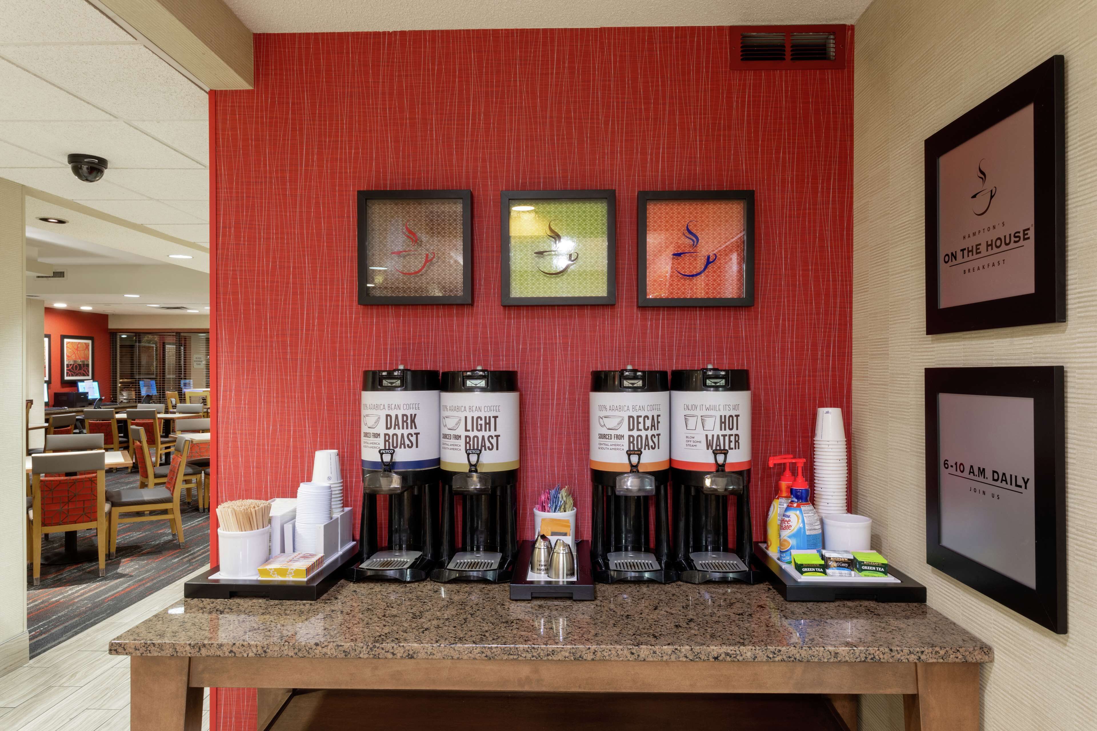 A coffee station with multiple coffee dispensers labeled 'Dark Roast,' 'Light Roast,' 'Decaf,' and 'Hot Water.' There are stacks of disposable cups and a tray with condiments and napkins.