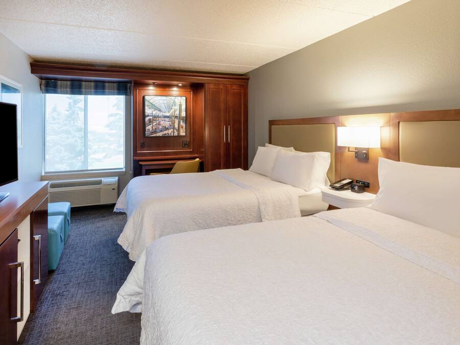 A hotel room with two beds, a desk, a television, and a window with a view of trees.