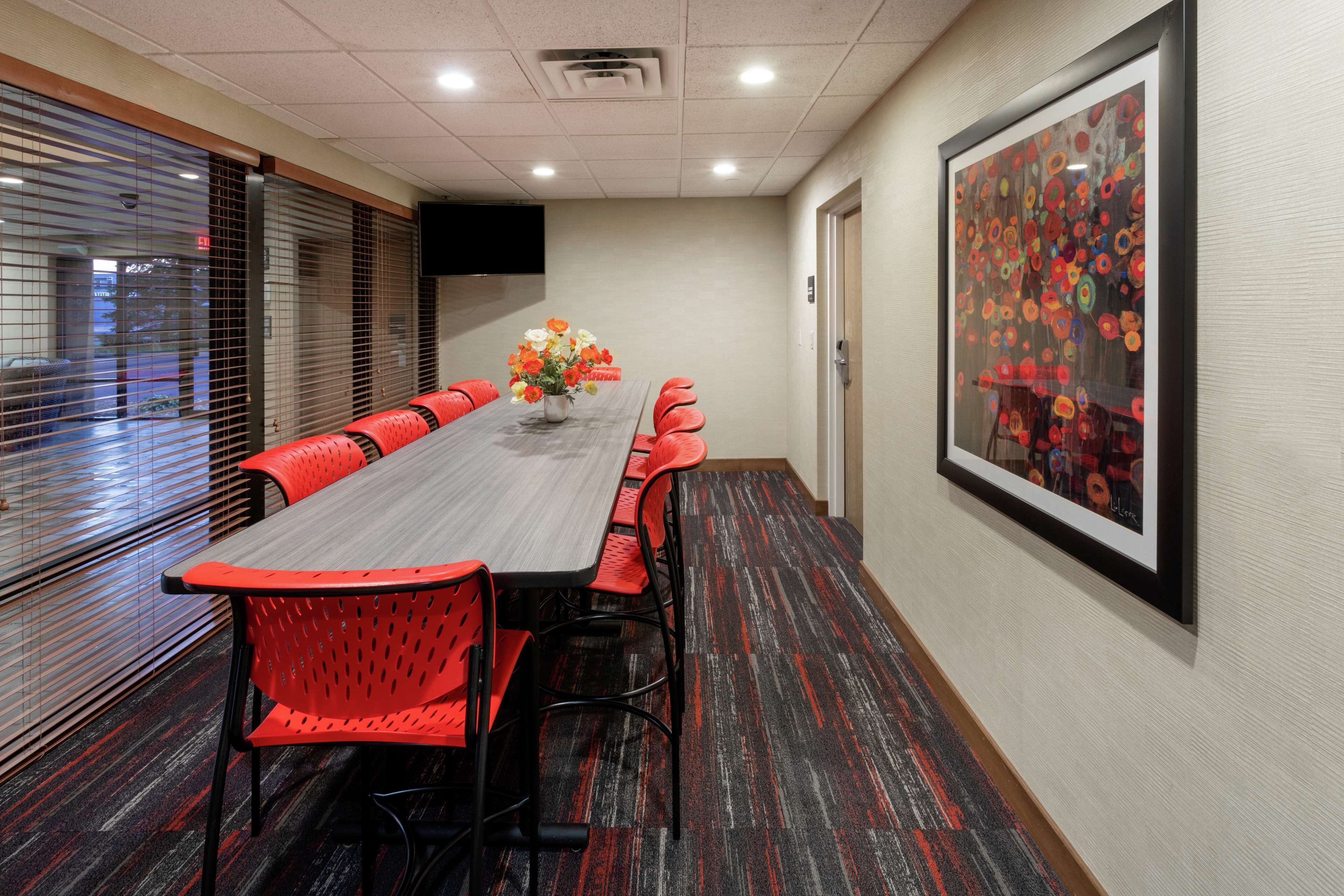 Un salón de conferencias con una larga mesa, sillas rojas, un jarr ón con flores y un gran cuadro abstracto en la pared.