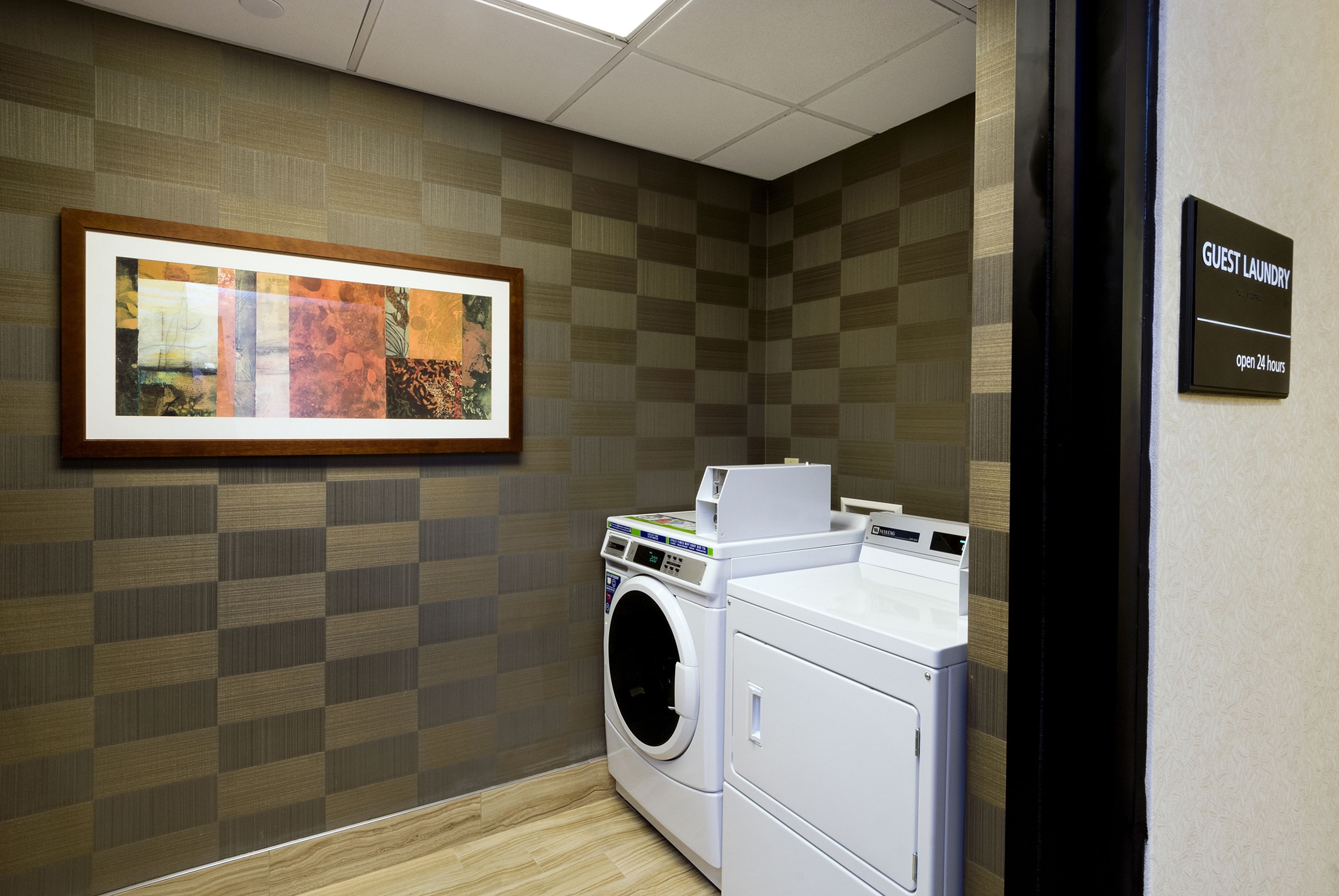 Une buanderie d’hôtel avec une grande œuvre d’art encadrée au mur, un lave-linge et un sèche-linge. Un panneau indique que la salle est ouverte 24 heures sur 24.