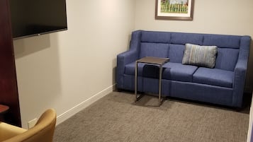 A room with a blue sofa, a small table, a TV mounted on the wall, and a carpeted floor.