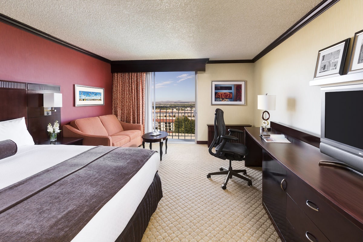 A hotel room with a large bed, a desk with a television, a chair, a small table, and a view of the city through the window.