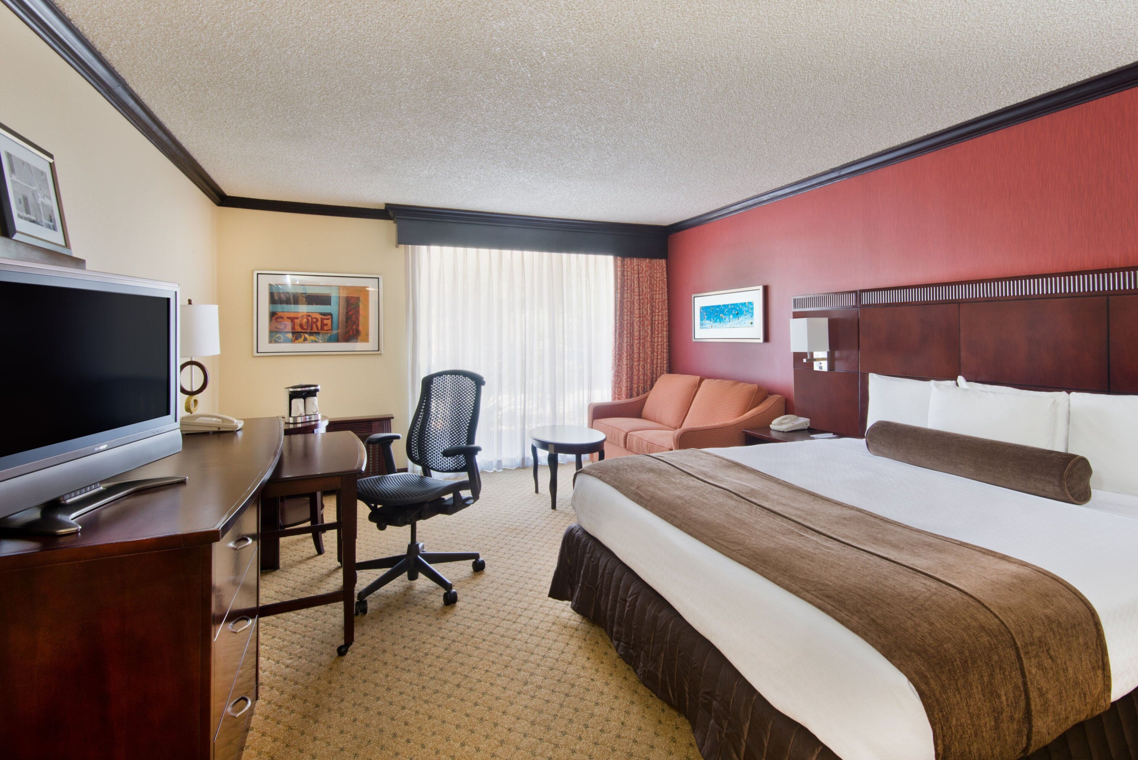 A hotel room with a bed, desk, chair, television, and artwork on the walls.