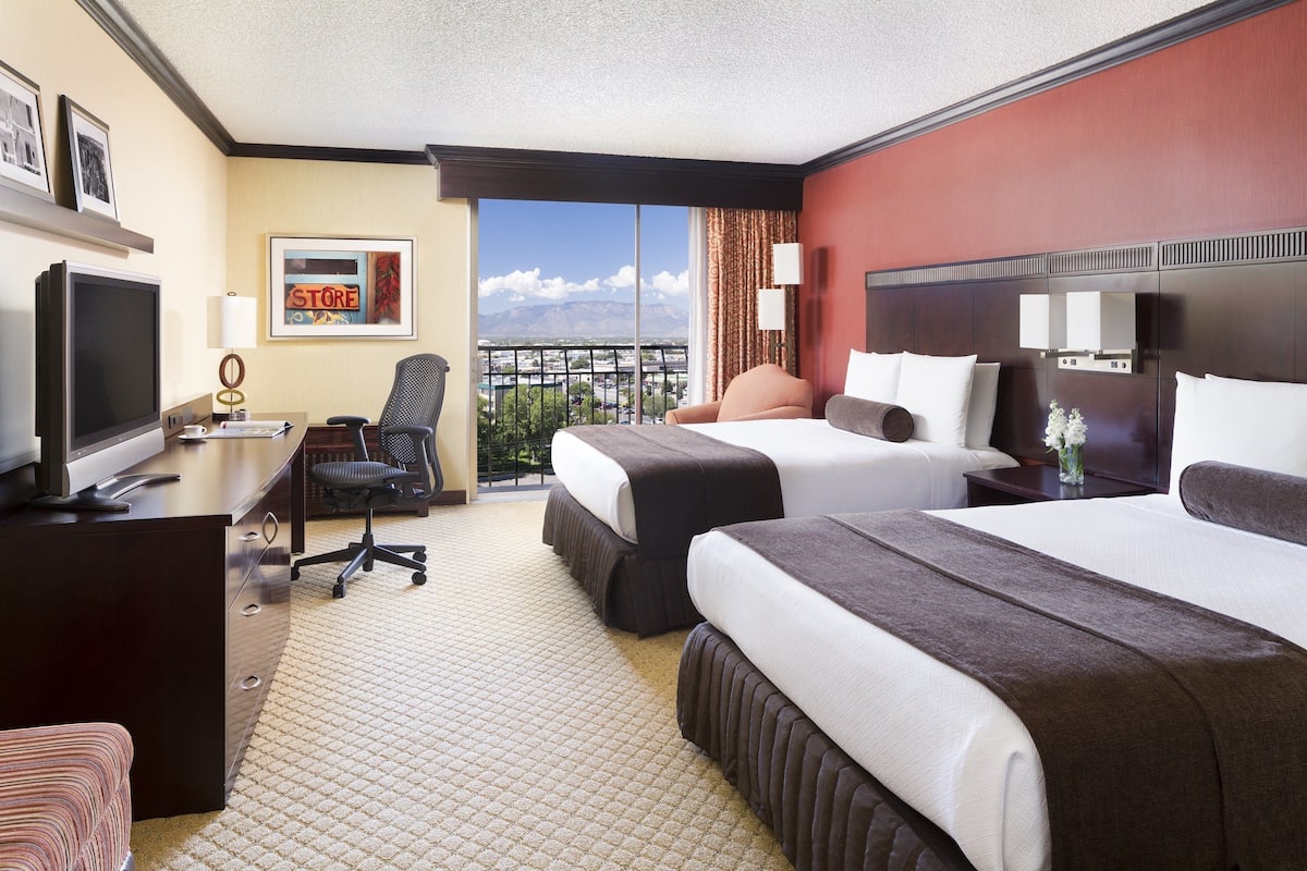 A hotel room with two beds, a desk with a TV, a chair, and a view of the city.
