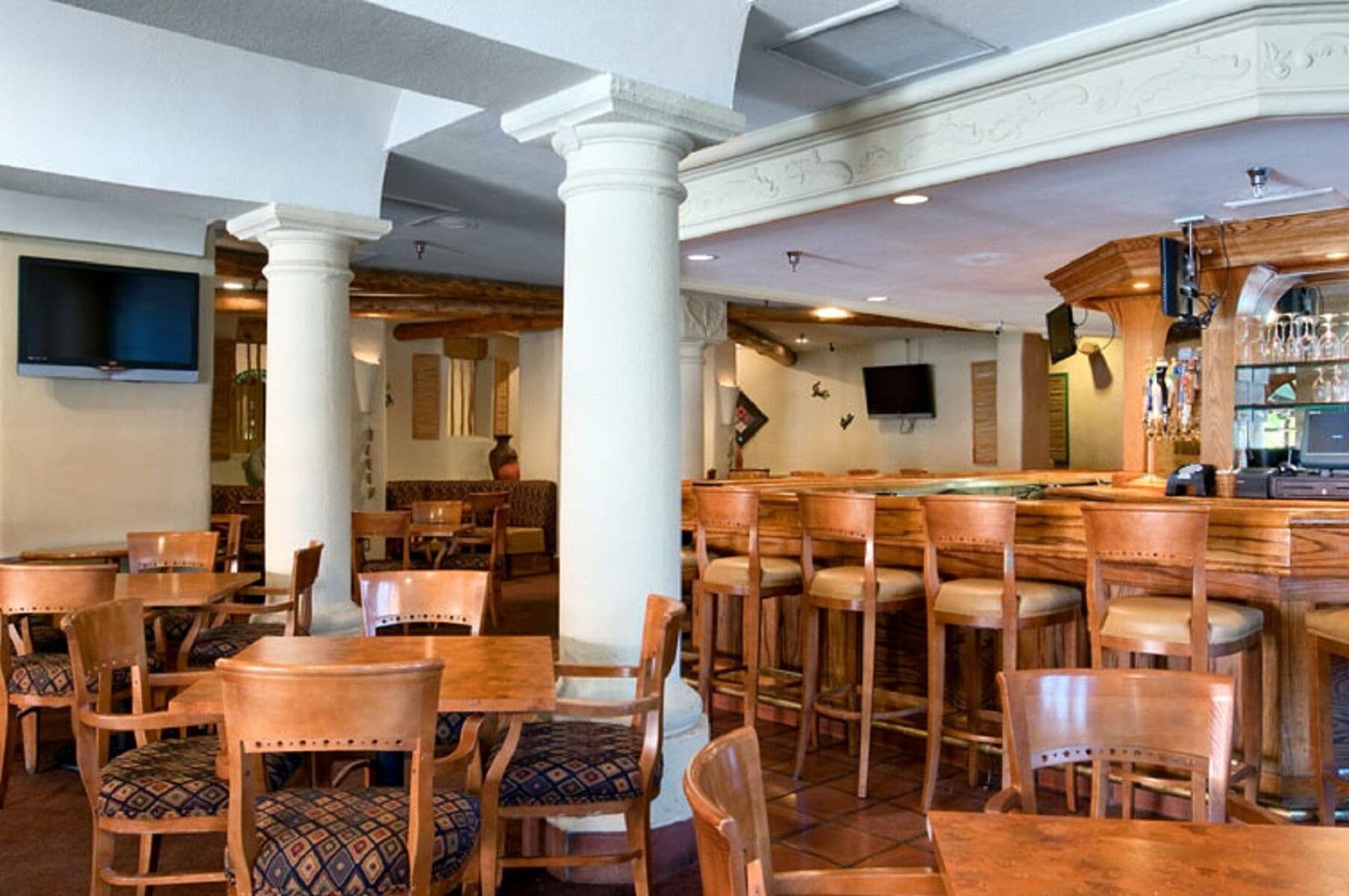 Zona de bar con mesas y sillas de madera, mesa de billar y barra con televisor y cristalería.