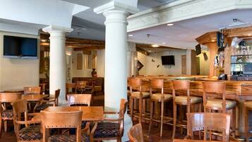 Zona de bar con mesas y sillas de madera, mesa de billar y barra con televisor y cristalería.