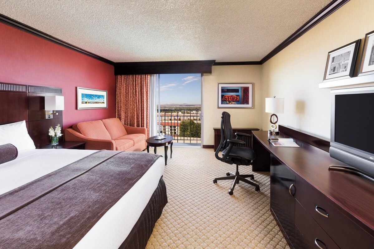 A hotel room with a large bed, a desk with a television, a chair, a small table, and a view of the city through the window.