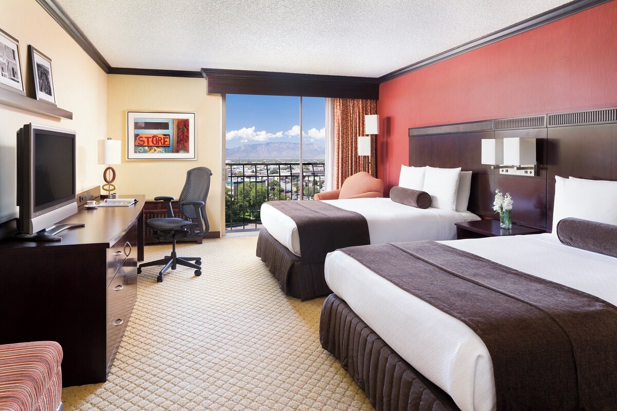 A hotel room with two beds, a desk with a TV, a chair, and a view of the city.