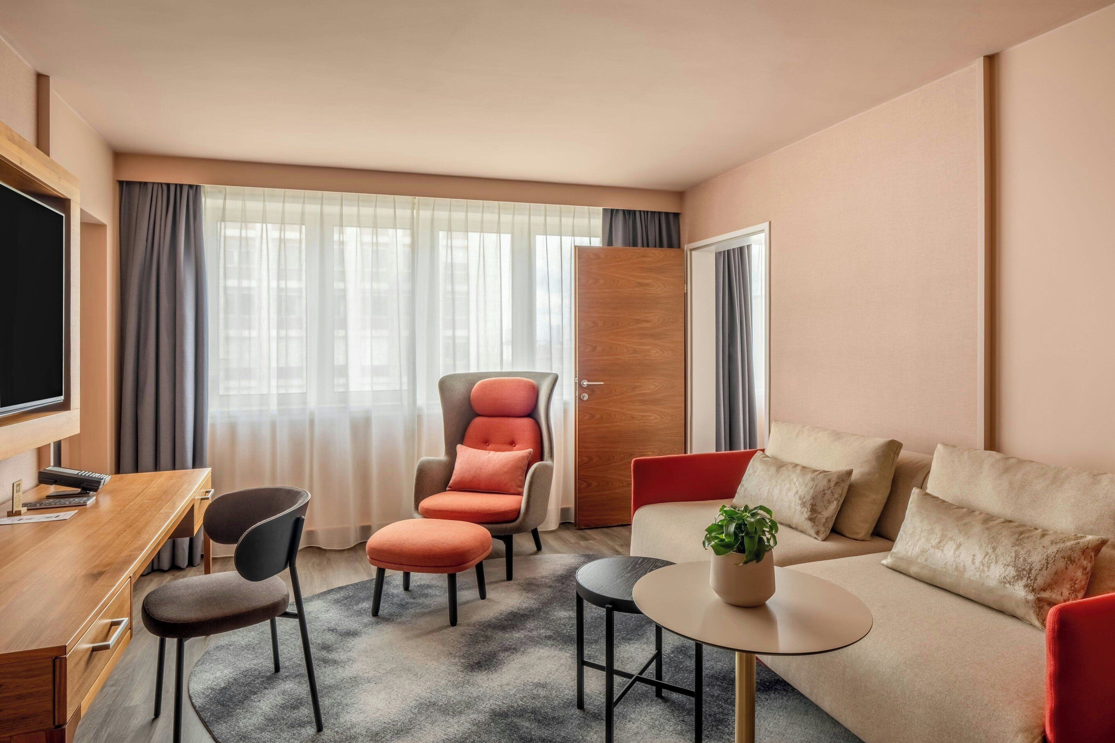 A modern hotel room with a wooden desk, a sofa, a red armchair, and a small round table with a plant.