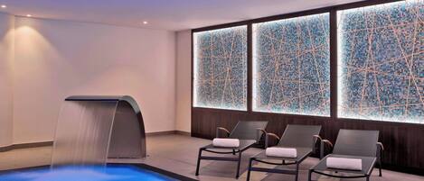 A modern indoor pool area with a water feature, lounge chairs, and mosaic-tiled walls.