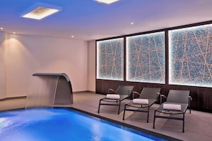 A modern indoor pool area with a water feature, lounge chairs, and mosaic-tiled walls.