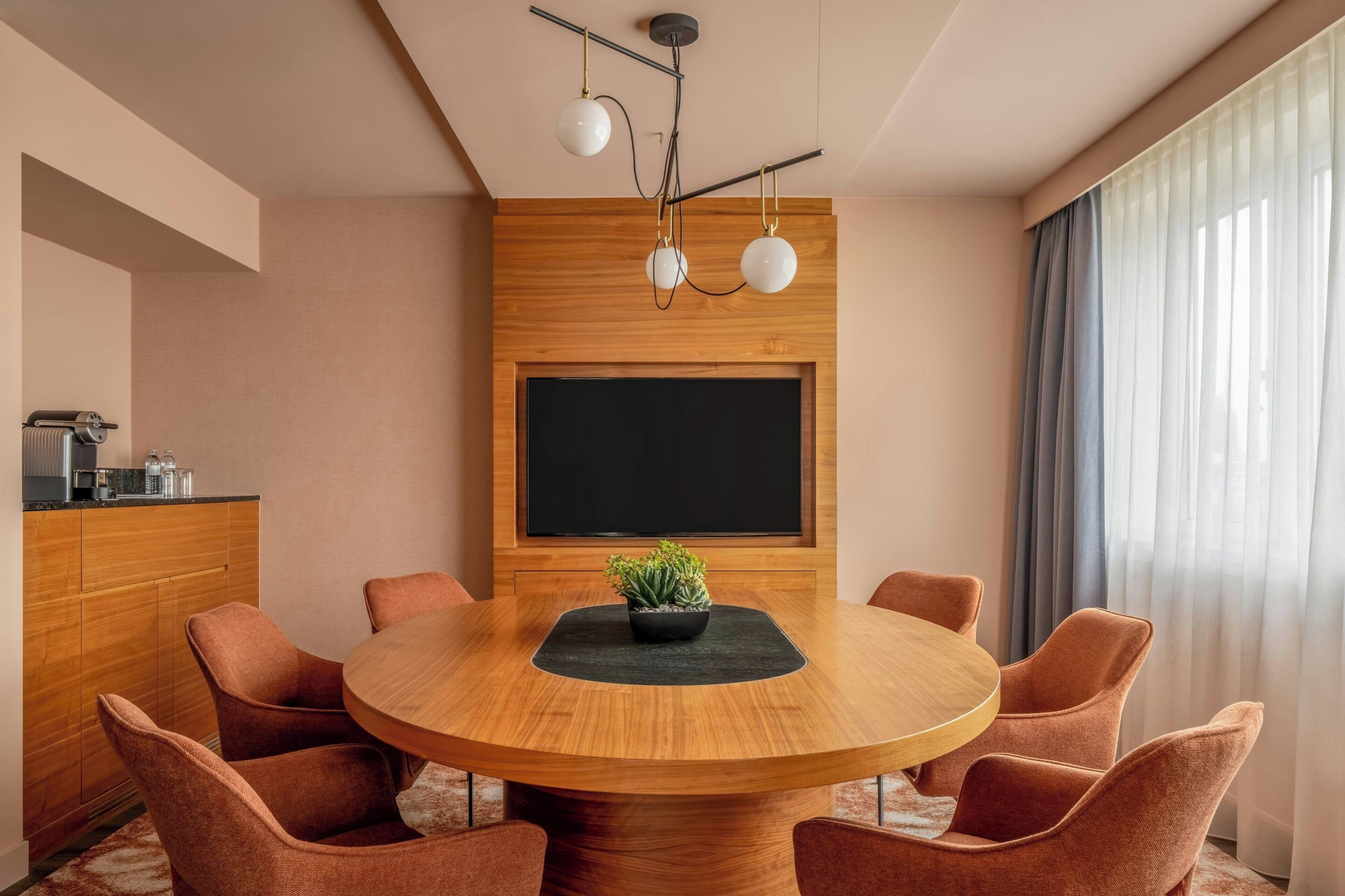 Ein moderner Essbereich mit einem runden Holztisch, sechs gepolsterten Stühlen, einem Blumentopf, einer Tafel und einer Küchenzeile mit Arbeitsplatte und Geräten.