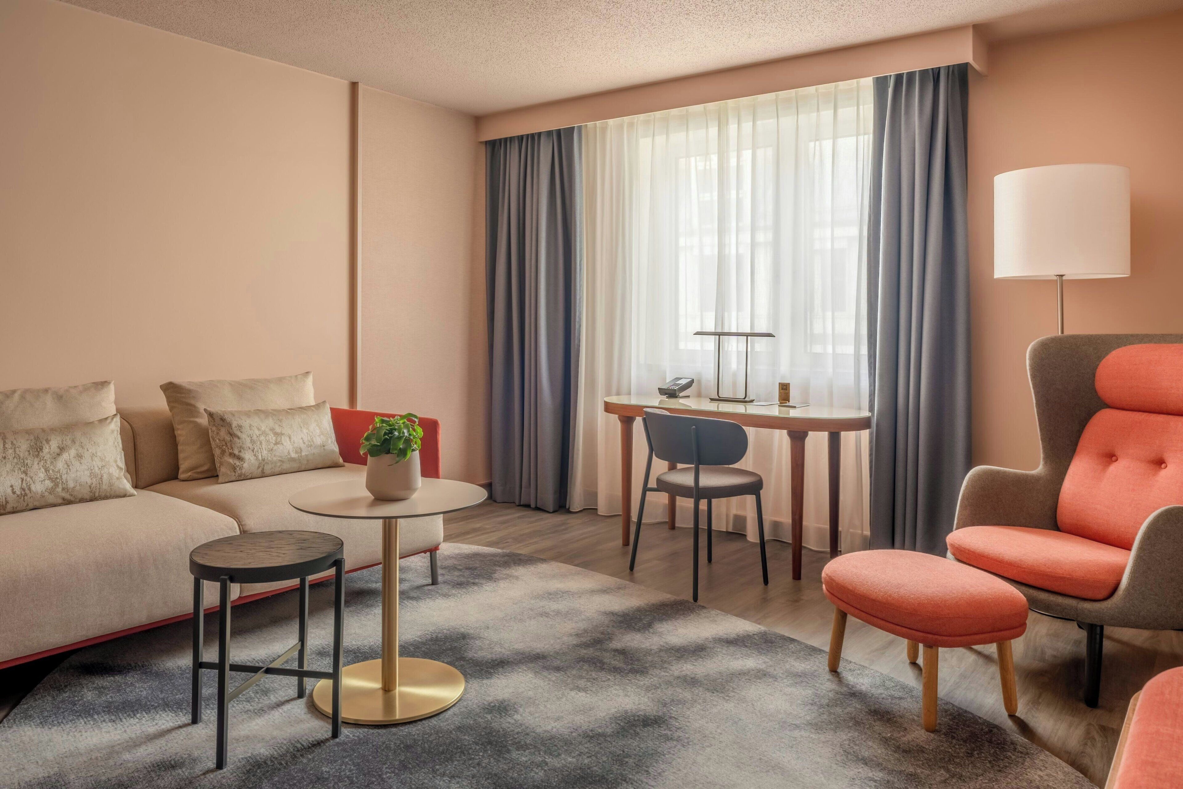 Quarto de hotel com sofá, poltrona, mesa redonda e uma pequena mesinha.
