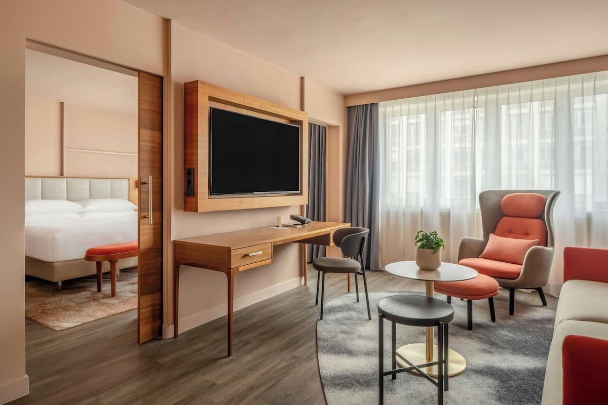 Moderni hotellihuone, jossa on sänky, työpöytä ja taso-tv, oleskelualue pyöreällä pöydällä ja tuoleilla sekä suuri ikkuna, jossa on verhot.