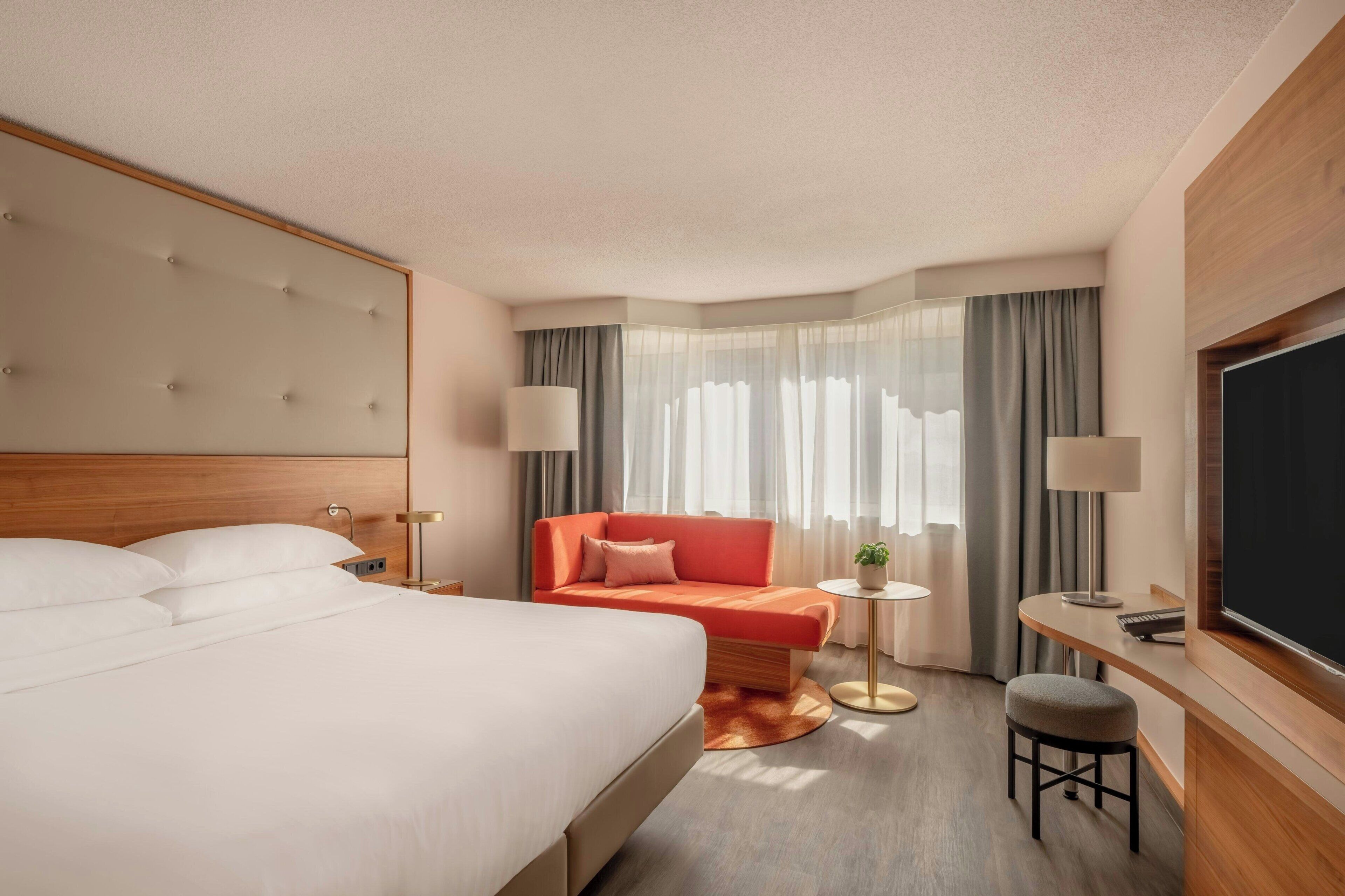 A hotel room with a large bed, a sofa, a small table, a TV, and a window with curtains.