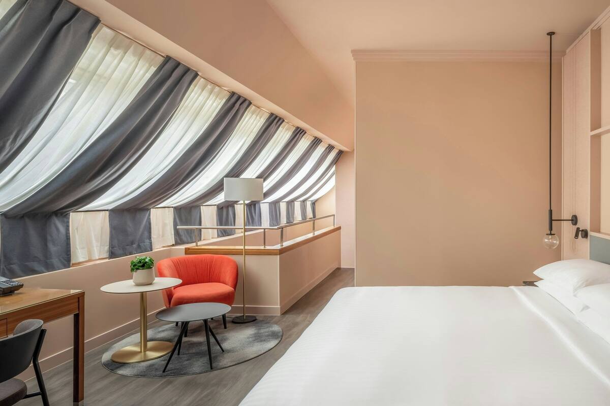 Moderni hotellihuone, jossa on suuri sänky, työpöytä tuolilla, pieni pöytä lampun kanssa ja tuoli. Huoneessa on ikkuna, jossa on verhot, ja ikkunasta on näkymä ulos.