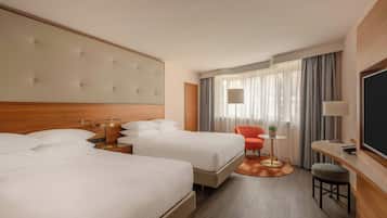 Quarto de hotel com duas camas, uma escrivaninha, uma cadeira, uma televisão e uma janela com cortinas.