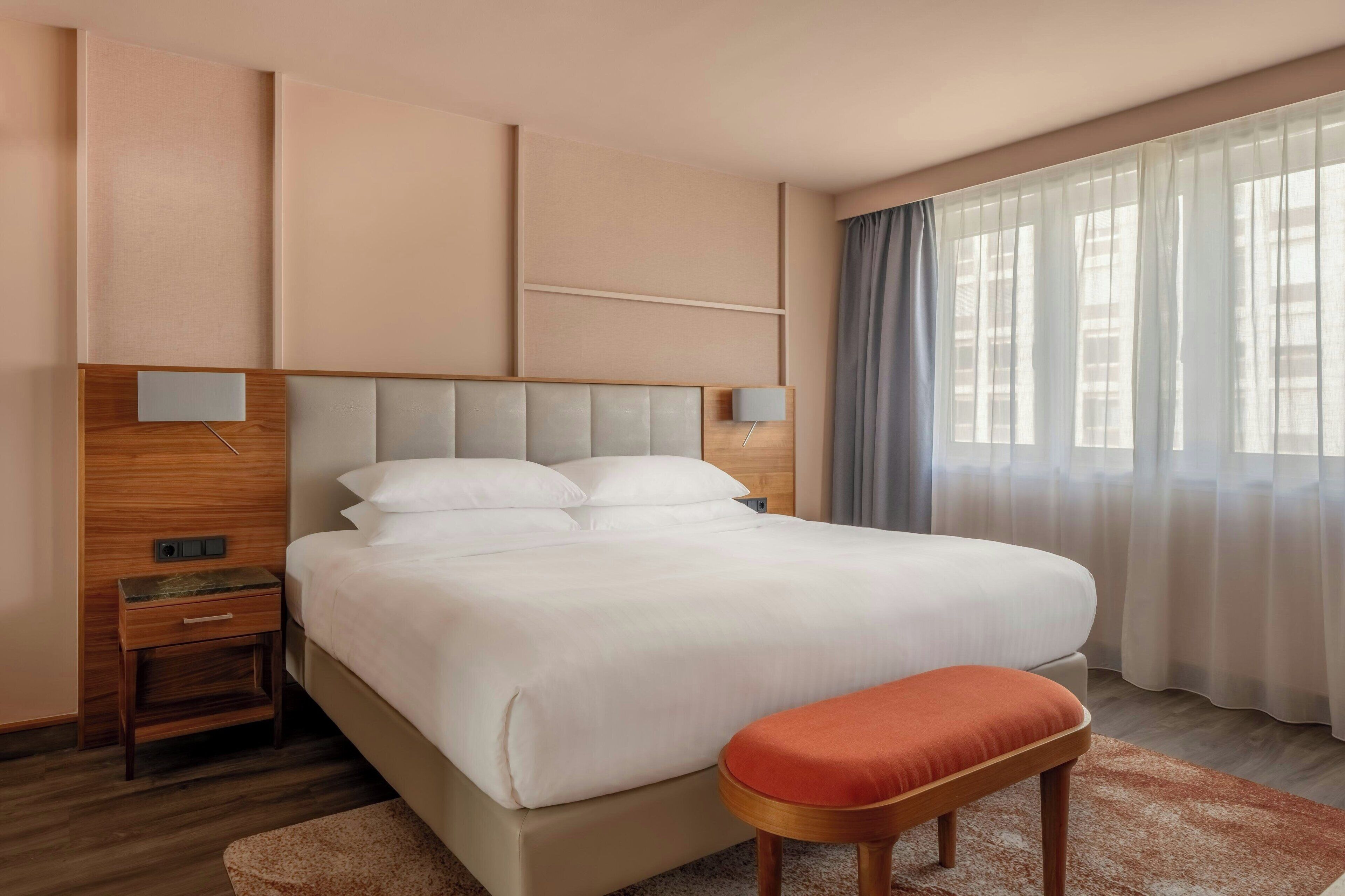 Quarto de hotel com uma cama grande, uma mesinha de cabeceira de madeira, um criado-mudo, um banco e uma janela com cortinas.