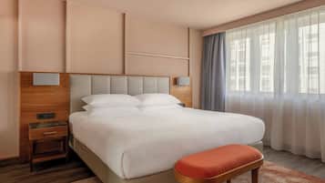 Quarto de hotel com uma cama grande, uma mesinha de cabeceira de madeira, um criado-mudo, um banco e uma janela com cortinas.