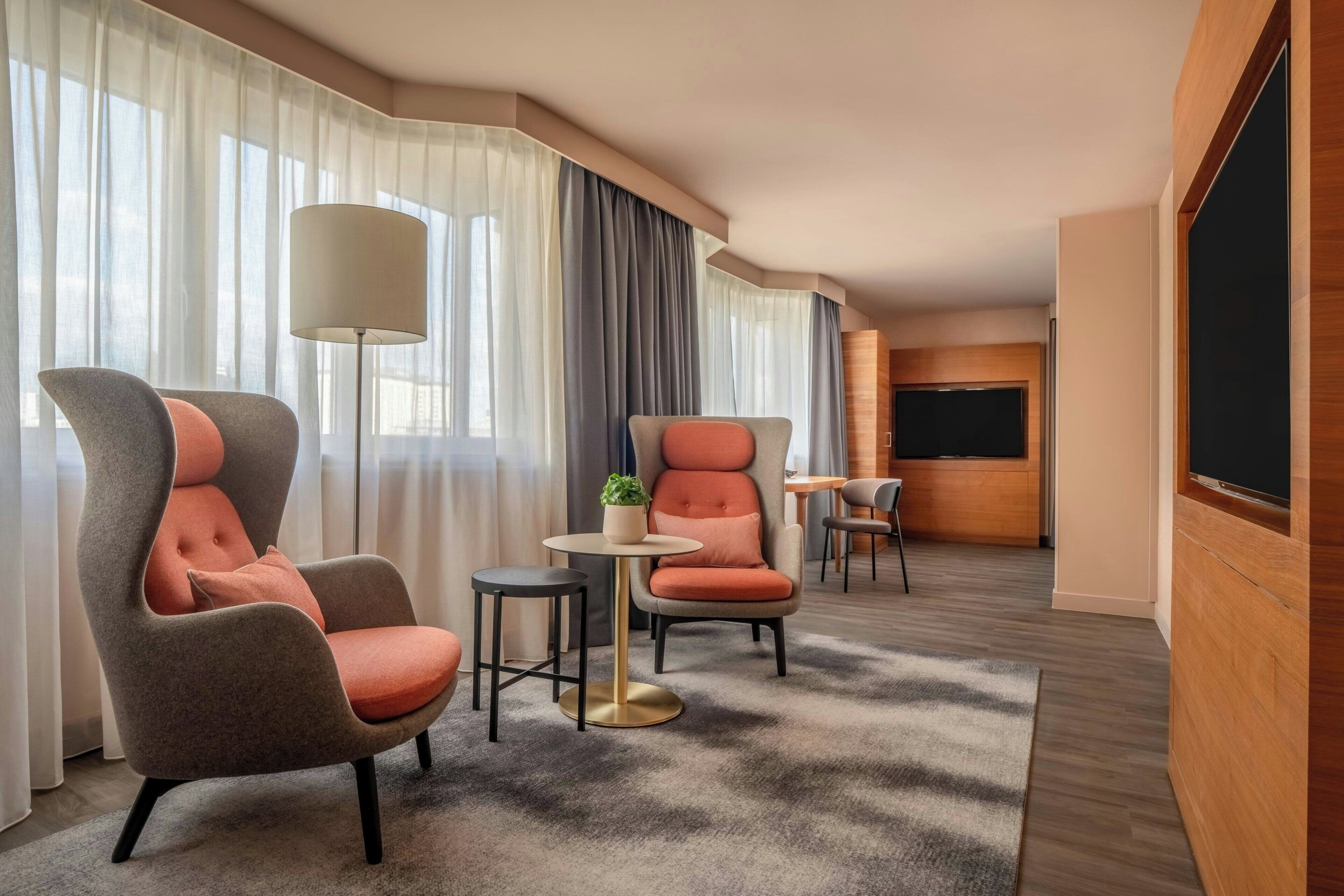A hotel room with two armchairs, a small round table, a floor lamp, a TV, and a large window with curtains.