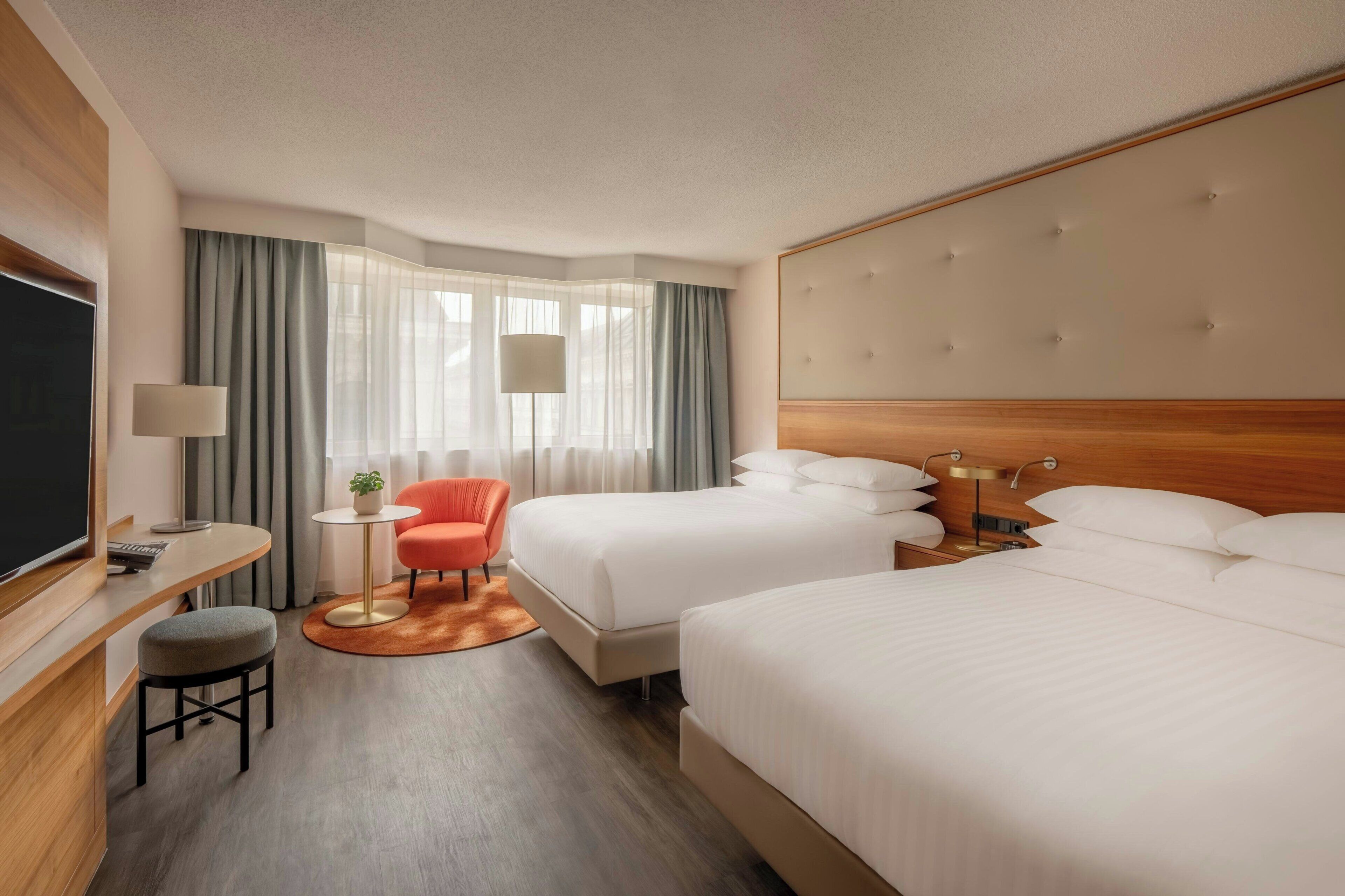 A hotel room with two beds, a desk with a chair, a small table with a plant, and a TV.