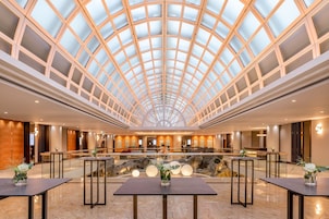 Um hall de hotel espaçoso com teto de vidro alto, um espelho d'água central e diversas áreas de estar.
