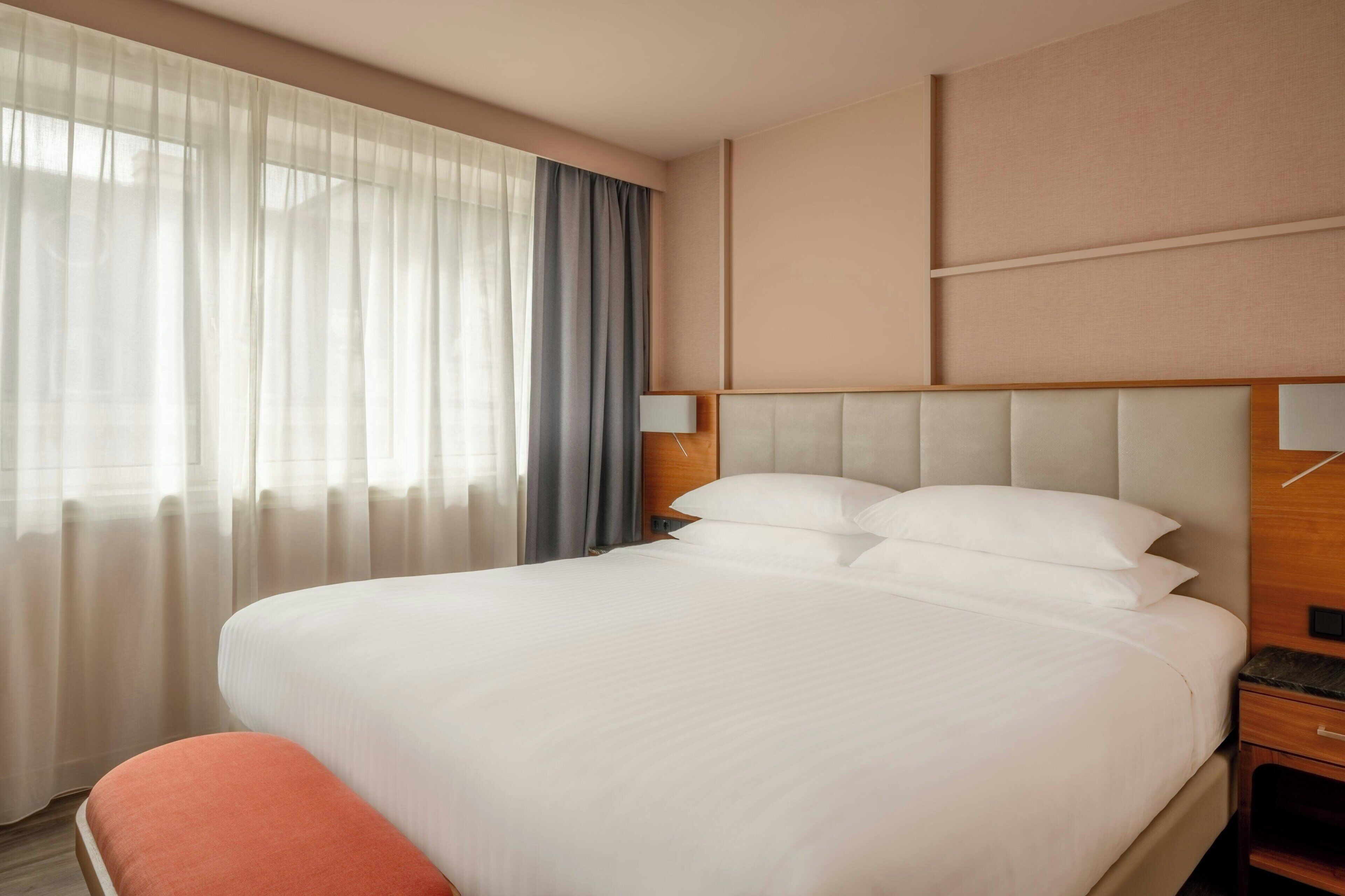 Habitación de hotel con una cama grande, una mesita de noche, cortinas translúcidas y una lámpara de pared.