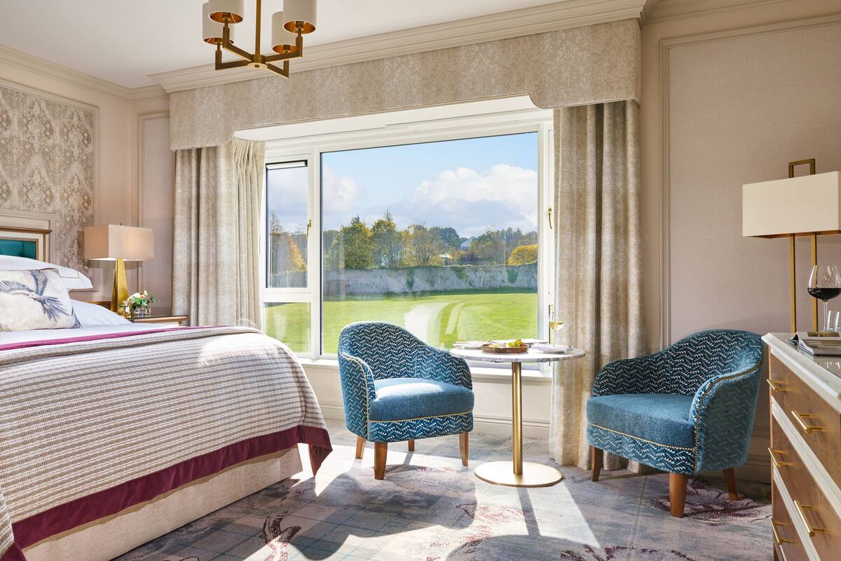 Ein Hotelzimmer mit einem großen Bett, zwei Sesseln, einem kleinen Tisch und Blick auf eine grüne Landschaft.