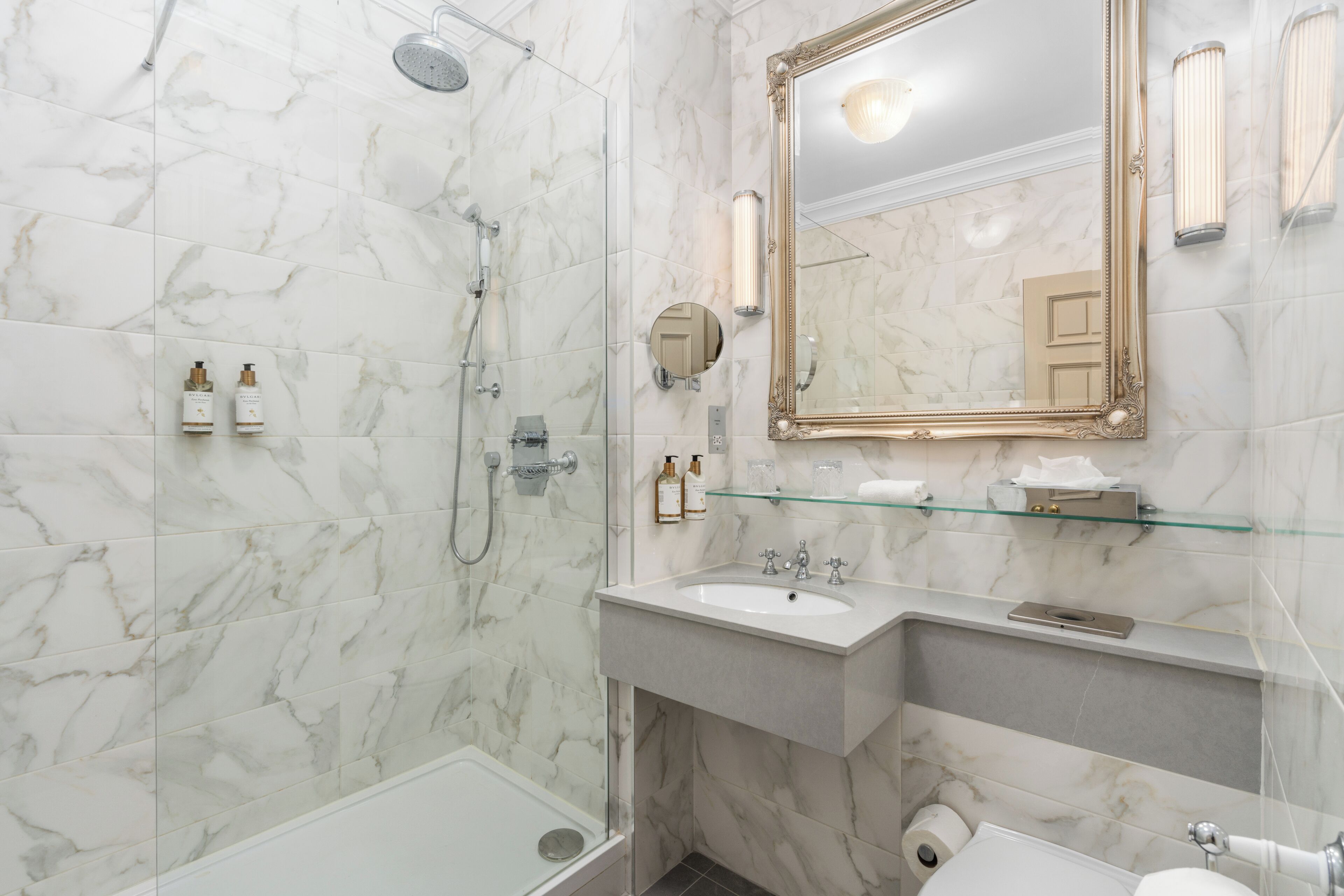 Un baño con suelo de mármol, mampara de vidrio, lavabo con espejo y artículos de aseo en la encimera.
