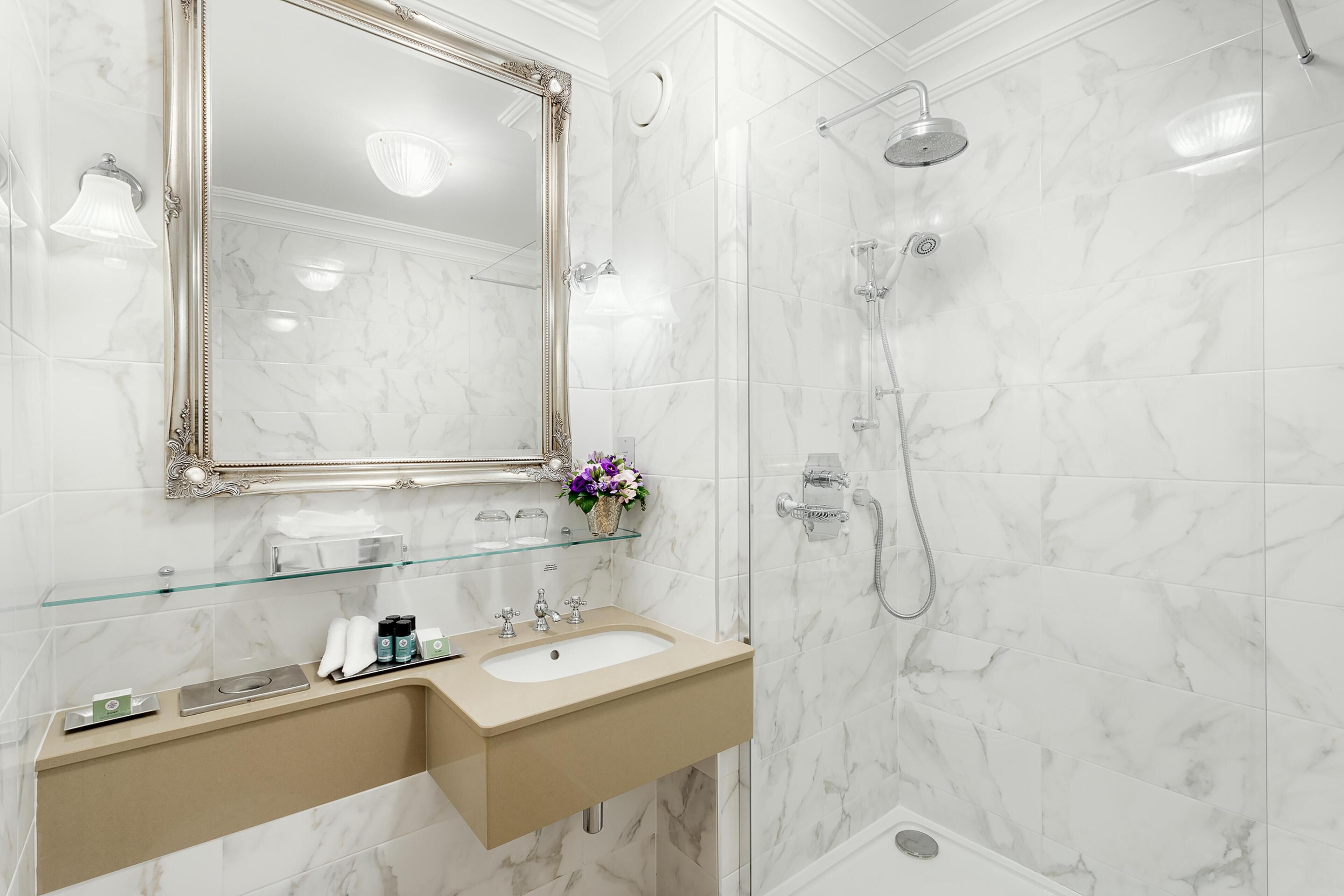 Ein Badezimmer mit Marmorwaschbecken, einer Glasablage für Toilettenartikel, einem großen Spiegel und einer Dusche mit Duschbrause.
