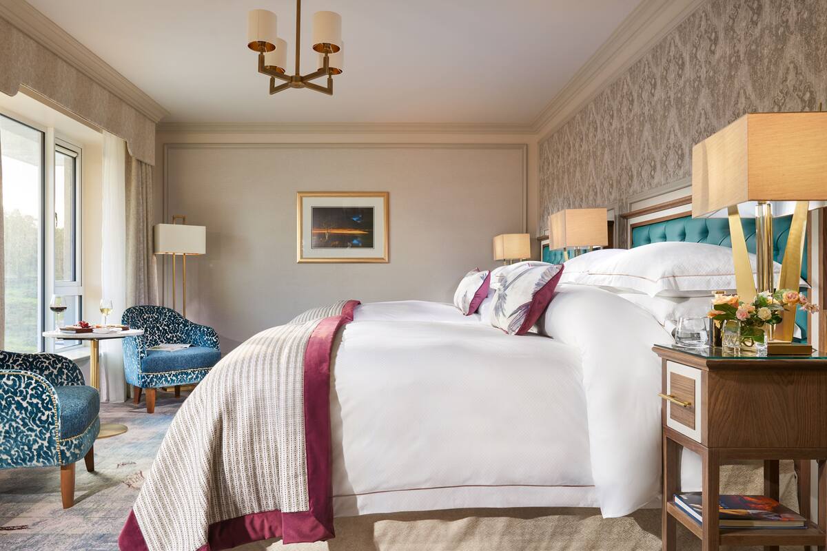 Ein Hotelzimmer mit einem großen Bett, einem Nachttisch mit Lampe, einem Sessel und Blick ins Freie.