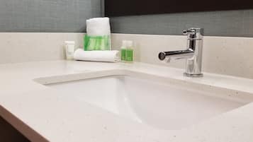 Ein Badezimmer mit einem weißen Waschbecken, einem Spiegel und Toilettenartikeln, darunter eine grüne Flasche, eine weiße Flasche und ein weißes Handtuch.