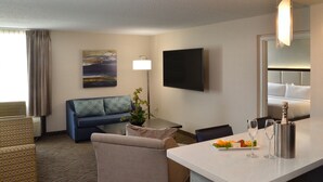A hotel room with a sofa, armchairs, a small table, a flat-screen TV, a lamp, and a minibar with a bottle and glasses. - Holiday Inn Youngstown South by IHG (Boardman)