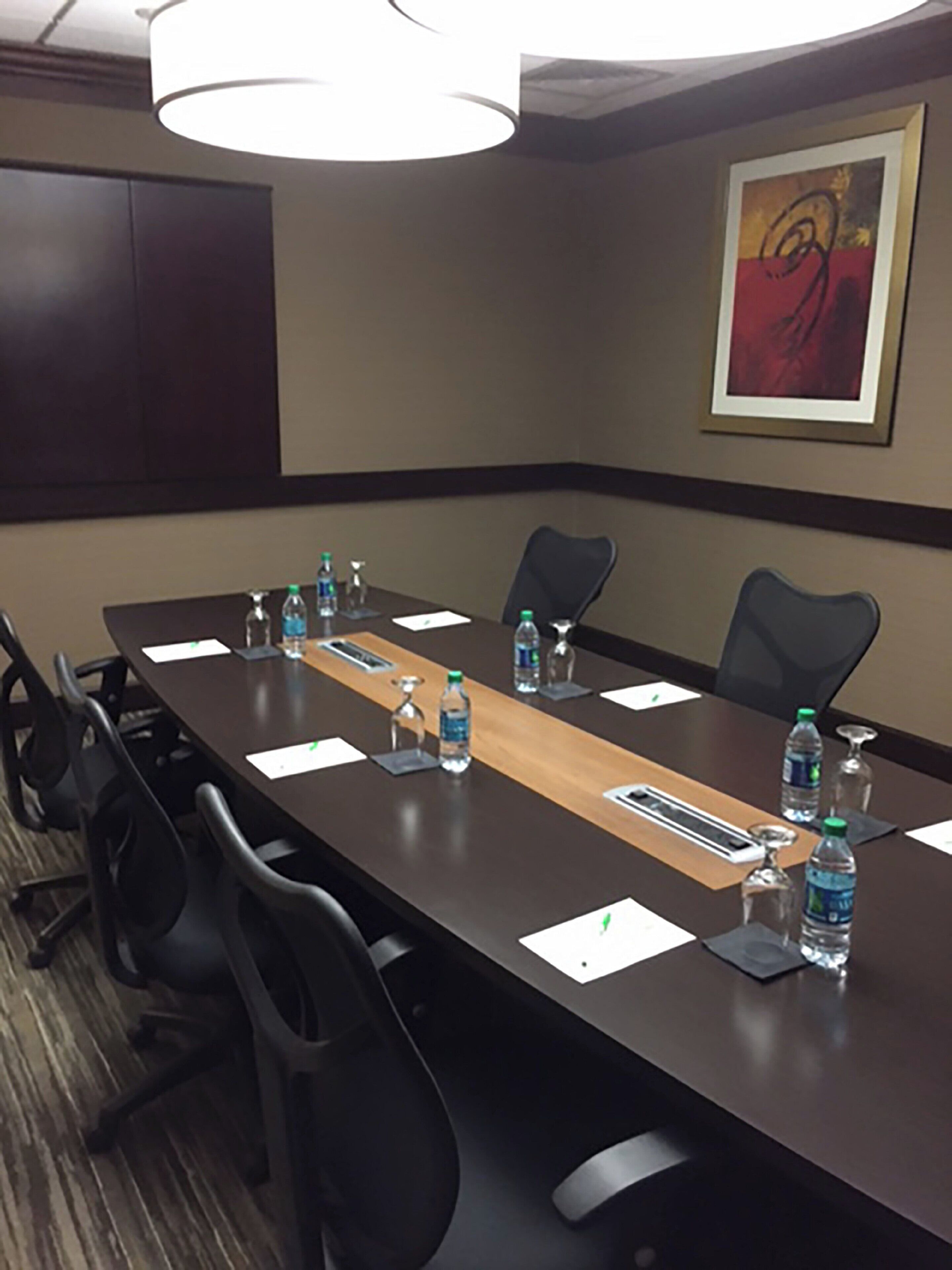 Sala de conferências com uma mesa comprida, cadeiras, garrafas de água e cartões de identificação, além de um quadro abstrato na parede.