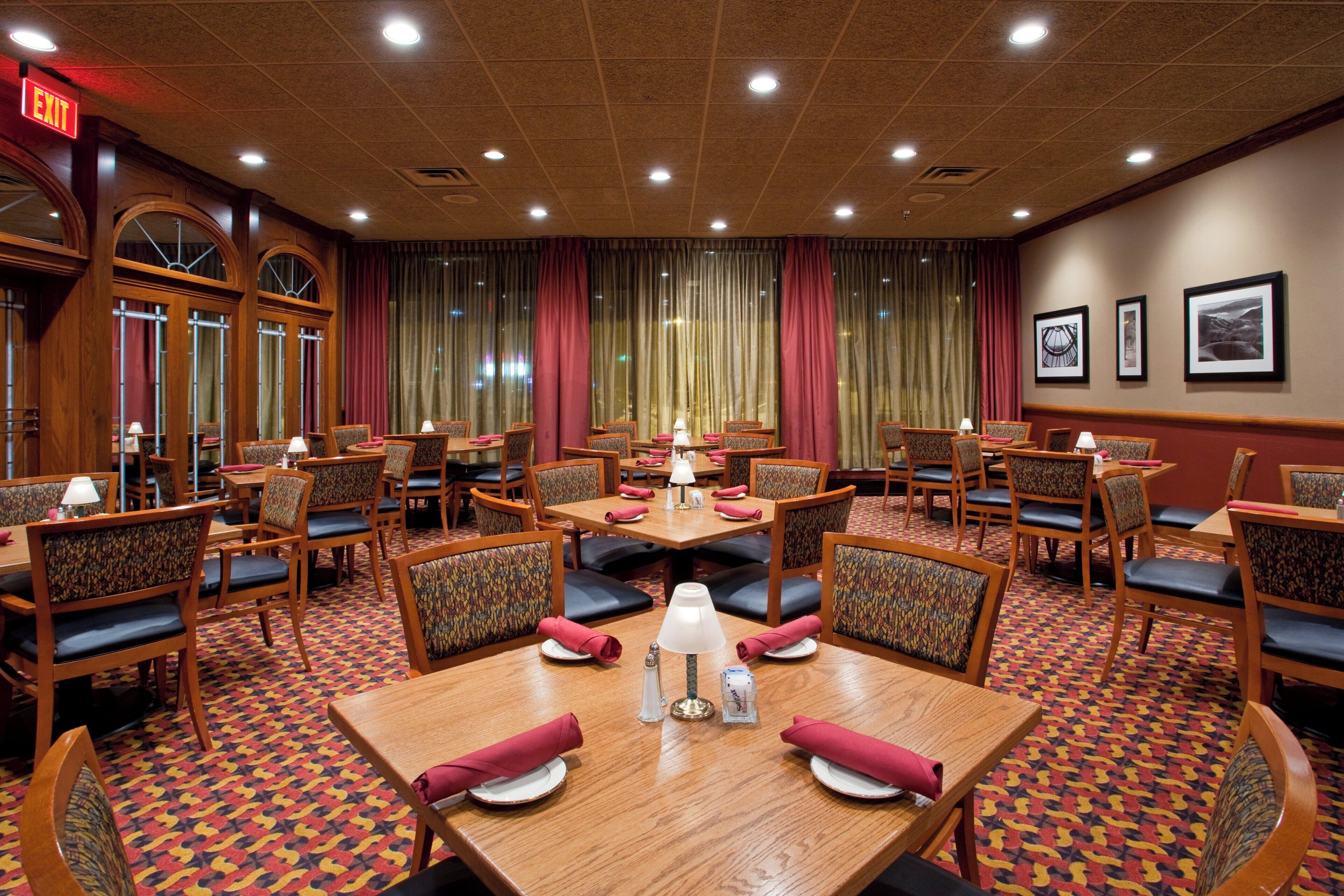 A hotel dining area with multiple tables and chairs, a patterned carpet, and a red exit sign.