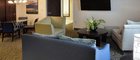 Habitación de hotel con sofá, sillón, mesa de comedor, sillas, televisor de pantalla plana y planta en maceta.