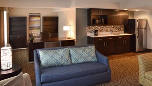 A modern kitchen with dark cabinetry, a stainless steel refrigerator, and a microwave. There is a blue sofa with patterned cushions in the living area. - Holiday Inn Youngstown South by IHG (Boardman)