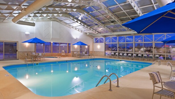 An indoor swimming pool with clear blue water, surrounded by lounge chairs and umbrellas, and large windows offering views of the outside. - Holiday Inn Youngstown South by IHG (Boardman)