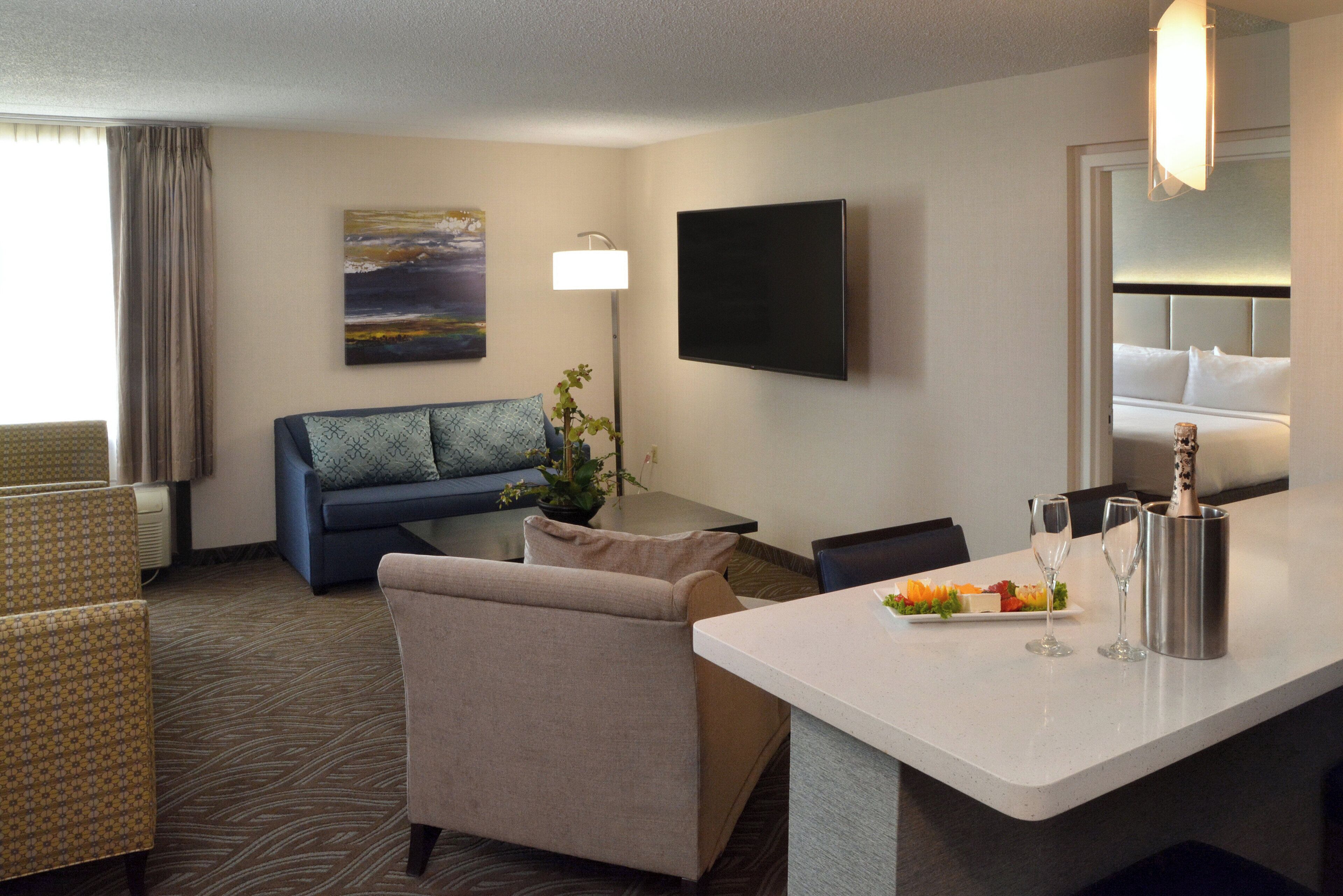 A hotel room with a sofa, armchairs, a flat-screen TV, a small table with a plant, and a breakfast bar with a plate of fruit and two glasses.